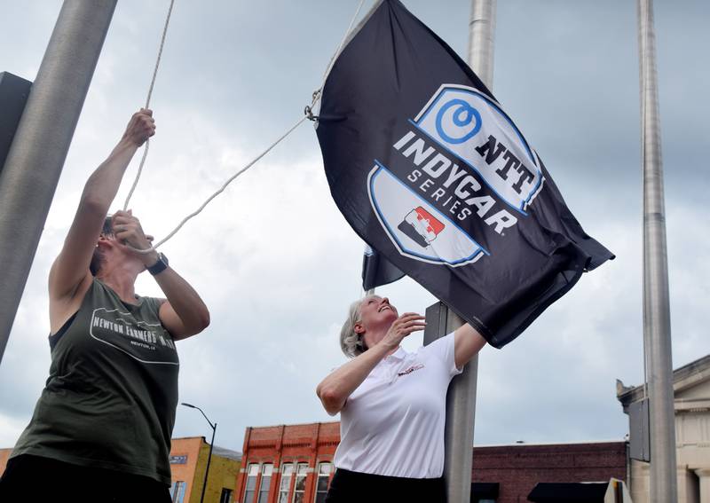 Erin Yeager, left, the executive director of Newton Main Street, prepares to hoist a flag to a post on June 25 in downtown Newton. She was joined by IndyCar representatives who helped place flags around the town square to commemorate the upcoming race weekend at Iowa Speedway.