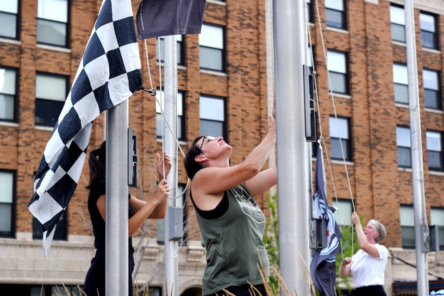 Erin Yeager, center, the executive director of Newton Main Street, prepares to hoist a flag to a post on June 25 in downtown Newton. She was joined by IndyCar representatives who helped place flags around the town square to commemorate the upcoming race weekend at Iowa Speedway.